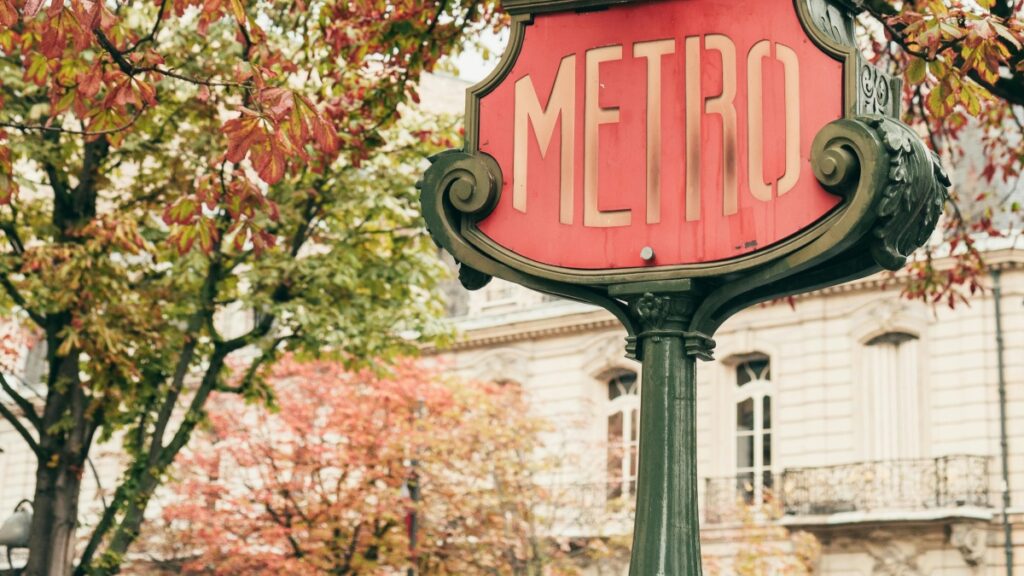 Un panneau rouge avec le mot "METRO" est affiché, emboîté dans un cadre décoratif en métal verdâtre. Dans le fond, il y a un bâtiment avec des fenêtres élégantes et des balcons, et des arbres aux feuilles automnales, avec des tons de rouge et de vert. En plus de cela, découvrez "Comment Gagner de l’Argent sur Triller : Guide Pratique pour les Créateurs en France" pour explorer les opportunités de gain sur les médias sociaux.