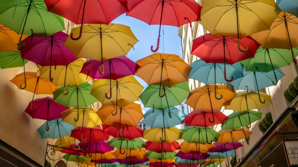 Un passage couvert orné de nombreux parapluies suspendus. Les parapluies sont de couleurs variées - rouges, verts, jaunes, oranges, bleus et violets - créant une mosaïque lumineuse. Le ciel bleu au - dessus ajoute à la beauté de la scène. Découvrez Comment Gagner de l'Argent sur WhatsApp Conseils Pratiques.