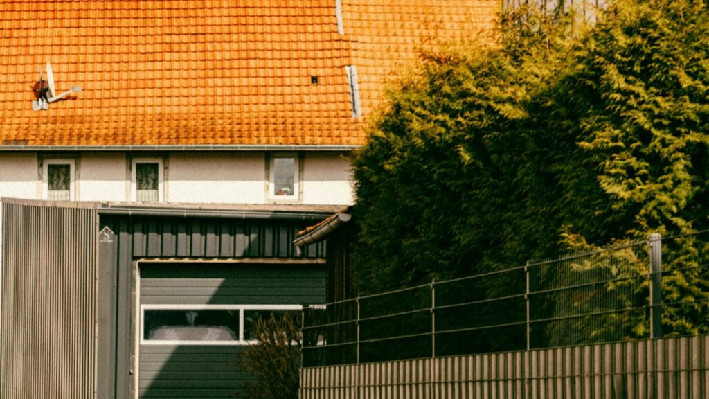 Une image d'un bâtiment avec un toit couvert de tuiles oranges et un mur gris. Il y a également une grande arbre verdoyant à côté du bâtiment et une clôture en métal en avant - plan. Découvrez Comment gagner de l'argent sur YouTube Guide pratique.