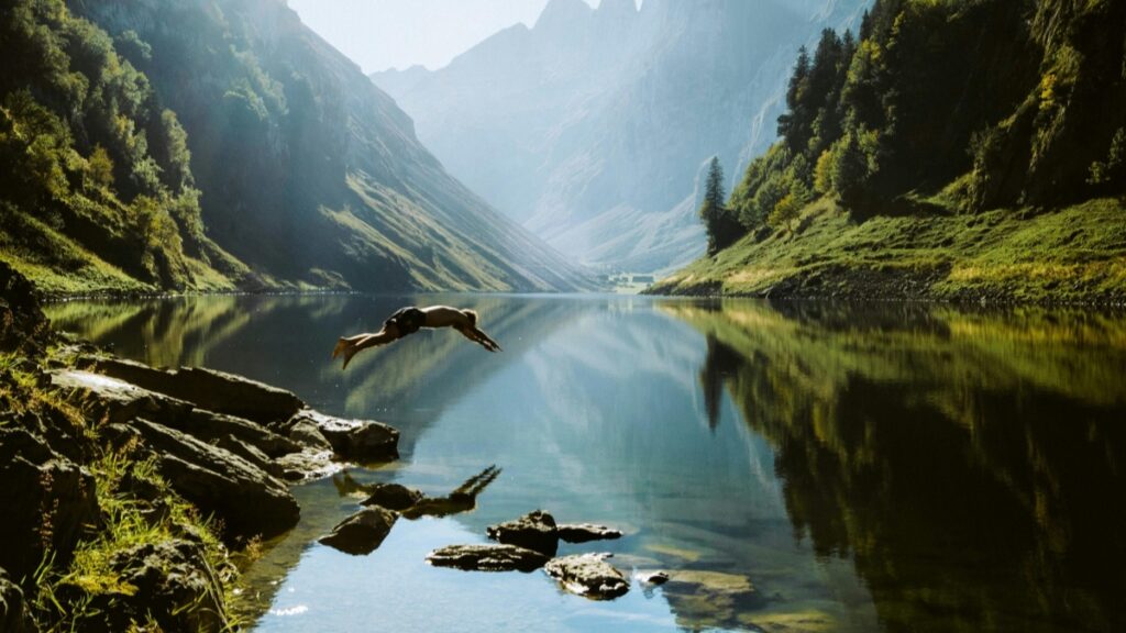 Una escena impresionante de un lago rodeado de montañas empinadas y cubiertas de verdor. La superficie del lago es tan calma que refleja perfectamente la belleza de los alrededores. Un individuo está en pleno salto hacia el agua desde la orilla, donde hay rocas y vegetación. Descubre más sobre Cómo Ganar Dinero a través de WhatsApp Consejos Prácticos.