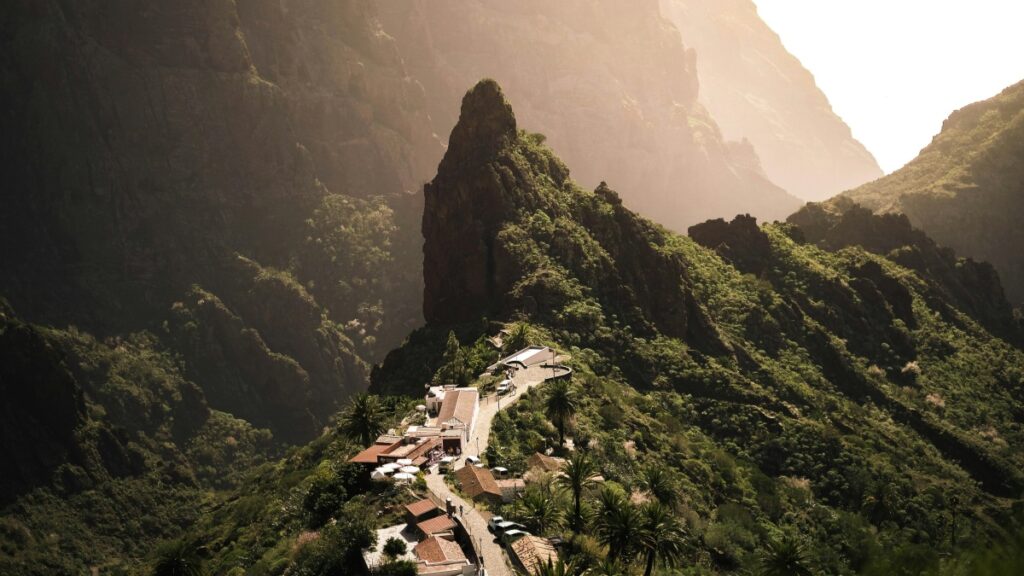 Una impresionante vista de un pueblo ubicado en una ladera de una montaña. Las montañas rodean el lugar, cubiertas de densa vegetación, con una roca prominente en el centro. El sol ilumina suavemente la escena, creando una atmósfera cálida. Además, descubre "Cómo Ganar Dinero en Telegram" para explorar nuevas formas de generar ingresos en las redes sociales.