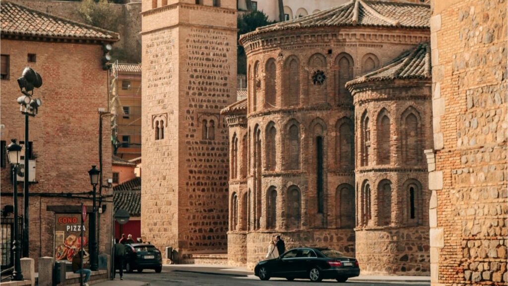 Esta imagen muestra un hermoso conjunto de edificios de estilo medieval, probablemente de origen religioso, con su característico uso de ladrillo y arcos. Las construcciones se encuentran en una calle con vehículos y un cartel de pizza, mostrando una mezcla de historia y vida cotidiana. Para aquellos españoles interesados en aprender a ganar dinero en Twitter, la guía "Cómo Ganar Dinero en Twitter: Guía Práctica para Españoles" ofrece valiosos conocimientos.