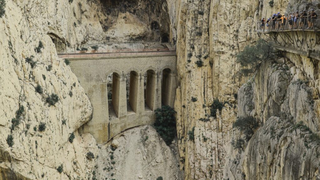 Esta imagen muestra un impresionante puente construido entre rocas en una garganta. El puente, con sus arcos y estructura sólida, se erige sobre un abismo, rodeado de rocas escarpadas. Además, hay un sendero suspendido en la roca donde un grupo de personas se encuentran. Si estás interesado en aprender a generar ingresos, no dejes de echar un vistazo a "Cómo Ganar Dinero en Vkontakte: Una Guía para Españoles", que te brindará valiosos consejos y estrategias.