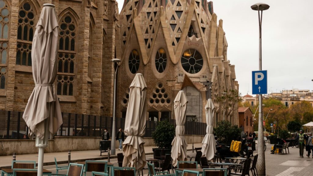 Una vista de una impresionante estructura arquitectónica, probablemente una iglesia o catedral, con un diseño único y detallado. Delante de ella, hay una zona de terraza con mesas y sillas, y paraguas plegados. También se ve un letrero de estacionamiento y algunas personas caminando por la calle. Descubre Cómo Ganar Dinero en YouTube Una Guía Práctica.