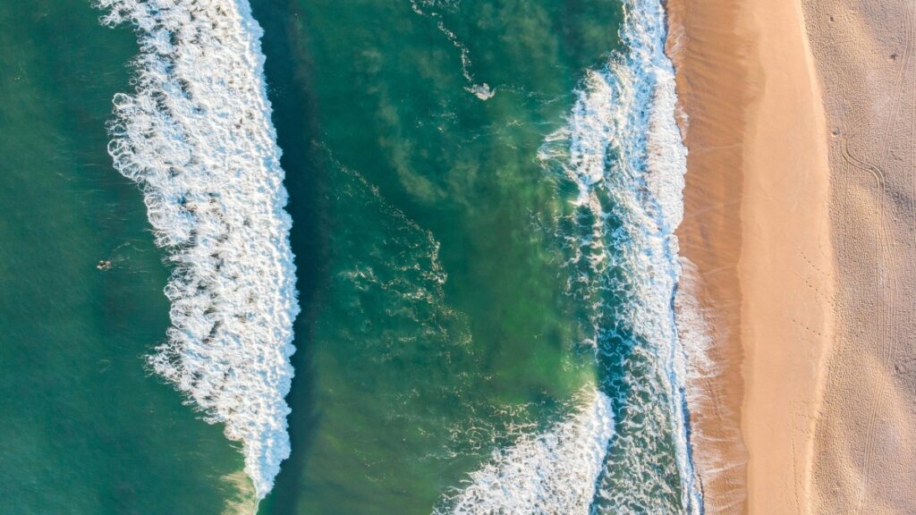 Vista aérea de uma praia, onde as ondas brancas se chocam contra as águas verdes - azuis do mar. A areia dourada forma um contorno suave ao longo da costa. A paisagem natural é impressionante, mostrando a força e a beleza da natureza. Além disso, descubra "Como Ganhar Dinheiro no Triller Um Guia Prático para Criadores no Brasil" e explore oportunidades de renda nas mídias sociais.