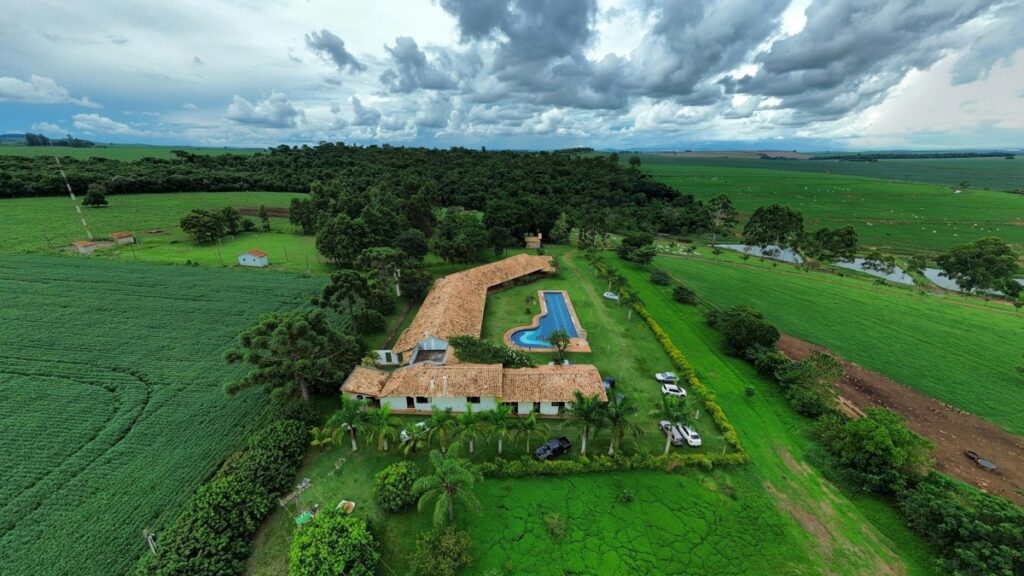 Uma vista aérea de uma propriedade rural, cercada por extensas plantações verdes e florestas. A propriedade inclui uma casa com telhado de barro, uma piscina azul e vários carros estacionados. As nuvens escuras no céu dão um contraste dramático à paisagem. Descubra Como Usar o Moj Lite para Marketing Empresarial no Brasil.