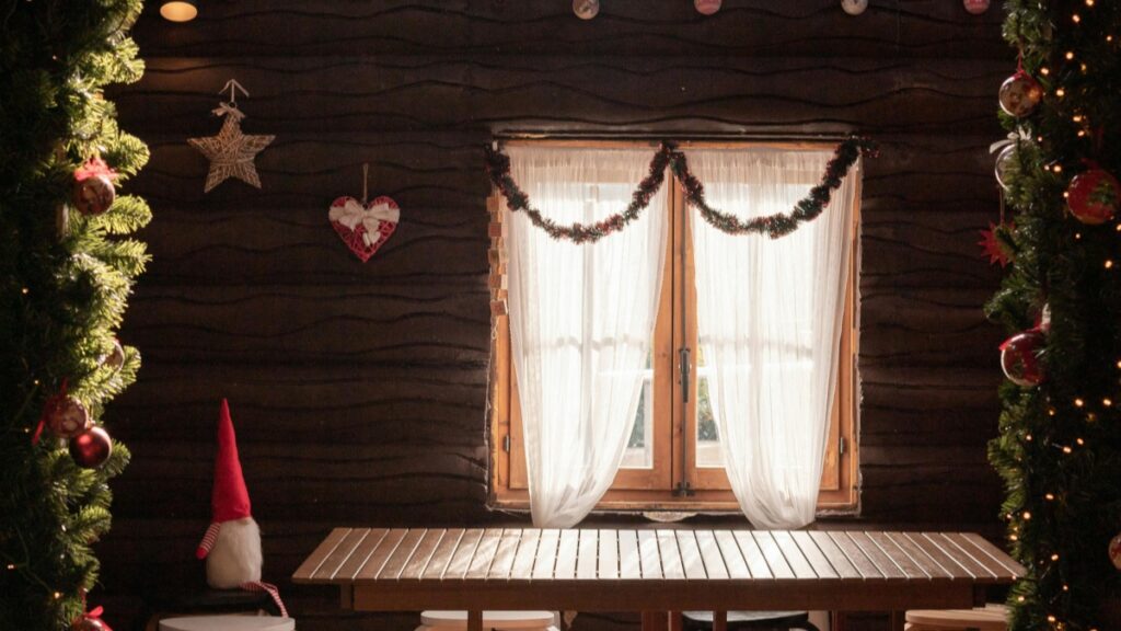 Interior de una estancia con un aspecto rústico, decorado con elementos navideños. Hay una ventana con cortinas ligeras, rodeada de guirnaldas. Las paredes de madera se adornan con adornos como estrellas y corazones. Además, hay un gnomo navideño y árboles de Navidad con luces y bolas. Aprende más sobre Cómo Utilizar WeChat para el Marketing Empresarial en España.
