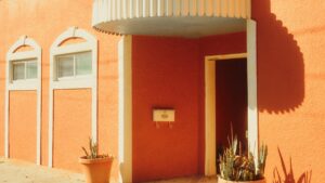 Das Bild zeigt das Fassade eines Hauses mit einer warm - orangefarbenen Außenwand. Zwei kleine Fenster mit weißen Rahmen und eine Eingangstür mit einem kleinen Dachvorsprung sind zu sehen. Ein Briefkasten befindet sich an der Wand neben der Tür. Zwei Pflanzkübel mit Pflanzen stehen vor der Tür. Erfahren Sie mehr über Geld Verdienen über WhatsApp Praktische Tipps.