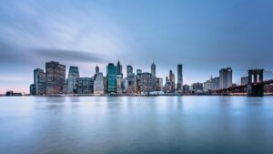 A stunning skyline of a major city at dusk, with a collection of modern skyscrapers reflecting off the calm water. The Brooklyn Bridge spans across the right side, adding an iconic touch. The sky is painted in soft hues of blue and pink. Discover more about How to Leverage Moj Lite for Business Marketing in the U.S.