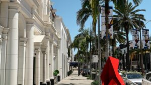 A sunny street scene lined with elegant white buildings featuring classical columns. Palm trees stand tall along the sidewalk, adding a touch of the tropics. Colorful banners adorn the poles, and a striking red modern sculpture catches the eye. As you soak in this vibrant urban atmosphere, also discover "How to Make Money on Triller" and unlock new income - generating opportunities in the world of social media.