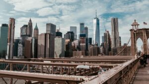 A striking view of a major city's skyline from a bridge, likely the Brooklyn Bridge. The foreground shows the bridge's metal framework and pedestrian walkway, while the background is filled with a dense cluster of skyscrapers, including the iconic One World Trade Center. The sky above is partly cloudy. Discover more about How to Make Money on YouTube A Practical Guide.