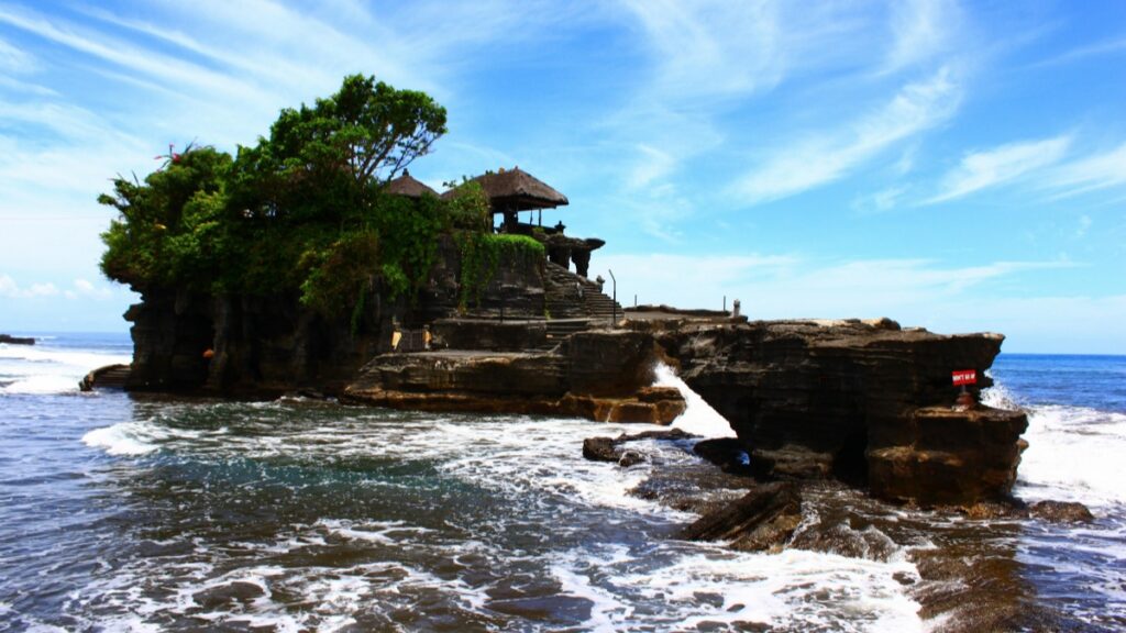 Pemandangan temple Tanah Lot di Bali, Indonesia, dengan ombak yang deras dan langit biru. Informasi tambahan: Standar Biaya Kerja Sama Influencer di ShareChat 2025 di Indonesia.