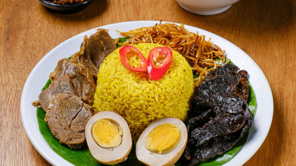 Piring makanan dengan nasi kuning yang dihiasi dengan cabai merah, ditambah daging empuk, telur rebus yang dipotong dua, serta rempah - rempah yang lezat. Makanan ini bisa menjadi inspirasi bagi konten influencer. Terkait dengan Standar Biaya Kerja Sama Influencer di Triller 2025 di Indonesia, presentasi makanan seperti ini bisa menjadi ide unggulan untuk menarik audiens.