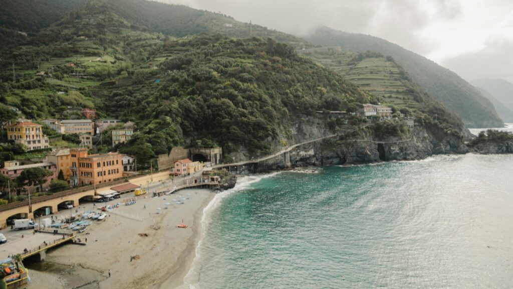Pemandangan pantai indah dengan ombak yang lembut, tanah pasir bersih, dan bangunan - bangunan berwarna di tepian pantai. Di belakang, gunung yang ditutupi dengan pepohonan menambah keindahan. Terkait dengan Standar Biaya Kerja Sama Influencer di Vigo Video 2025 di Indonesia, lokasi seperti ini bisa menjadi latar belakang yang hebat untuk konten kreatif yang menarik perhatian audiens.