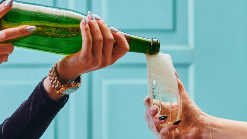 Deux mains, l'une portant une montre et avec des ongles peints en blanc, l'autre avec des ongles peints en noir, versant une boisson pétillante d'une bouteille verte dans un verre. La boisson déborde en mousse. Cette image est associée aux Tarifs des Collaborations avec des Influenceurs sur Zalo en France en 2025.