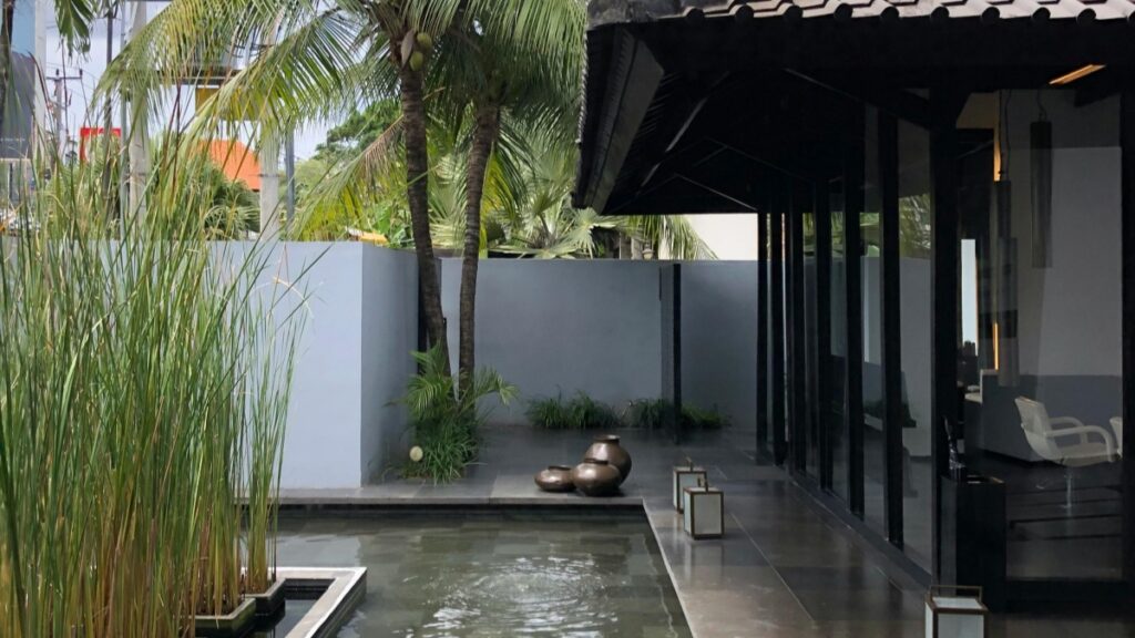 Gambar ini menunjukkan sebuah area luar ruangan yang tenang, dengan taman air dan pohon - pohon, seperti pohon kelapa. Desain arsitektur modern dan minimalis terlihat pada bangunan di sampingnya, dengan dinding gelap dan jendela - jendela besar. Bagi Anda yang ingin mengetahui lebih lanjut tentang platform video alternatif, kunjungi "Vigo Video Saudara TikTok yang Terlupakan" untuk mendapatkan wawasan yang berharga.