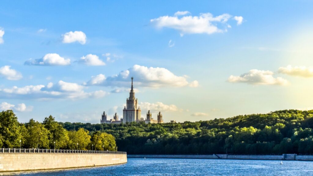 Вид на Moscow State University с реки. Городской пейзаж сочетает в себе мощный исторический комплекс с высокой башней и зелёный лес, который окружают его. Небо расцветает в синие тона с белыми облаками, а солнце светит ярко. И не забудьте изучить "Как Заработать Деньги на Triller Практическое Руководство для Создателей в России", чтобы узнать о возможностях заработка в социальных медиа.