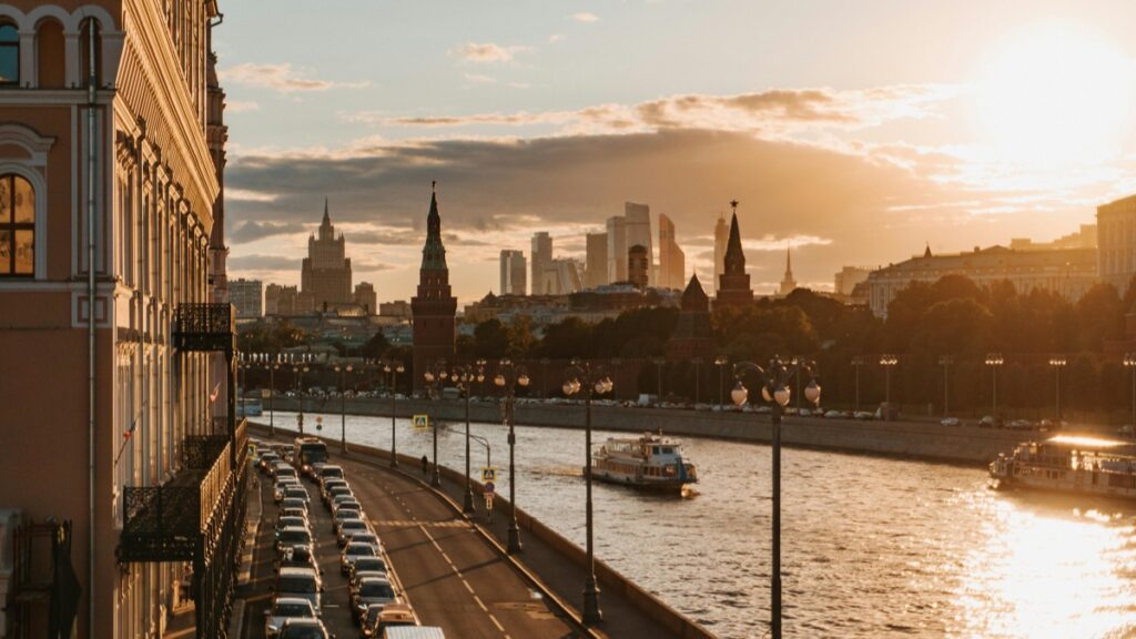 Вид на городской пейзаж во время заката. На берегу реки стоит ряд автомобилей, а на воде плывут паромы. В фоне silhuettes современных зданий и исторических сооружений с острыми шпилями. Узнайте, Как заработать деньги на YouTube Практическое руководство.