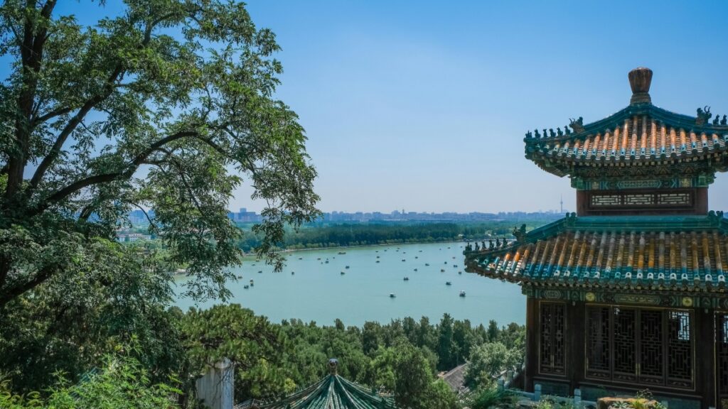 画面展现中式古典建筑一角，飞檐翘角，琉璃瓦色彩斑斓，尽显传统建筑之美。建筑旁绿树成荫，郁郁葱葱。远处是开阔的湖面，湖面上点缀着许多小船，湖岸与城市天际线相连，蓝天澄澈。在欣赏这美景的同时，不妨了解 “如何在 Triller 上赚钱”，探索社交平台新的创收途径。
