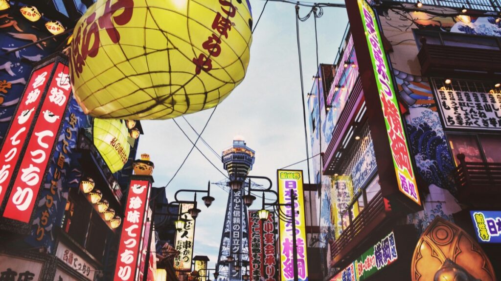 にぎやかな日本の街並み。華やかな商店の看板と提灯が並び、独特の雰囲気を漂わせています。背景には目を引くタワーがそびえ立ちます。2025年日本Kwaiインフルエンサー推奨協力料金基準に関する情報も、この活気ある街の風景と共に、日本のメディアやビジネスの側面を反映しています。